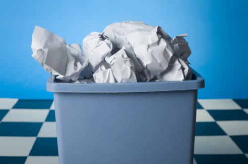 A close-up view of a rectangular, light blue plastic rubbish bin filled with crumpled white newspaper and paper waste, situated on a checkered black and white tiled floor. The background features a plain, bright blue wall, creating a vibrant contrast with the bin and waste material. The crumpled papers are disorderly arranged, spilling slightly over the top edge of the bin. The surface of the bin appears smooth with a matte finish, and the lighting highlights the creases and textures of the crumpled paper. The setting suggests an indoor space, possibly a kitchen or utility room, where waste is collected for removal or recycling. Queens Park House Clearance services often handle similar rubbish disposal tasks in the London area, including nearby postal districts, ensuring efficient management of household waste.