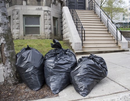 Three large black plastic rubbish bags filled with waste are placed on the sidewalk near the base of a tree, adjacent to a paved area. The bags are slightly shiny, bulging with contents, and tied at the top. Behind them, there is a stone building with a small, rectangular window and a set of external stairs with metal handrails leading up to an entrance. The scene is set in an urban environment, possibly in Queens Park, with a concrete pavement and a grassy patch visible behind the tree. The overall setting suggests a typical rubbish removal scenario in a residential or commercial area, with Queens Park House Clearance providing local waste disposal services visible through the context.