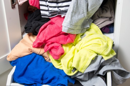 A close-up view of an open white storage drawer filled with a variety of crumpled and folded clothing items, including bright yellow, pink, blue, grey, black, and beige fabrics. The clothing appears casual and mixed, with some pieces made of soft, lightweight materials such as cotton or jersey, identifiable by their textures and finishes. The drawer is part of a larger storage unit situated outdoors or in a designated waste or disposal area, with a glimpse of a paved surface visible on the right side of the image. The scene suggests a cluttered area intended for rubbish or clothing clearance, possibly part of a house clearance service carried out by Queens Park House Clearance, in or around the London borough of Queen's Park. The overall environment indicates an organized effort to remove and dispose of unwanted textiles and garments. Lighting is natural, emphasizing the vividness of the clothing colors and the textures of the fabrics.