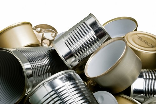 A collection of various metal tin cans, including both opened and unopened items, stacked and piled together. The cans are primarily in silver, gold, and metallic tones, with some showing ridged or smooth surfaces and others with pull-tab lids. They are arranged closely on a flat surface, likely outdoors on a driveway or pavement, with some cans leaning or partially overlapping each other. The background is minimal, emphasizing the metallic textures and reflective surfaces of the cans, indicative of waste materials awaiting collection or disposal. This scene illustrates typical household rubbish, which Queens Park House Clearance may handle in their house and waste clearance services in the London area.