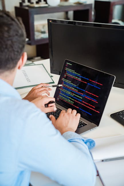 A person working at a desk with multiple monitors and a laptop displaying lines of computer code. The individual is using a keyboard and holding a pen, with their back partially visible. The desk also