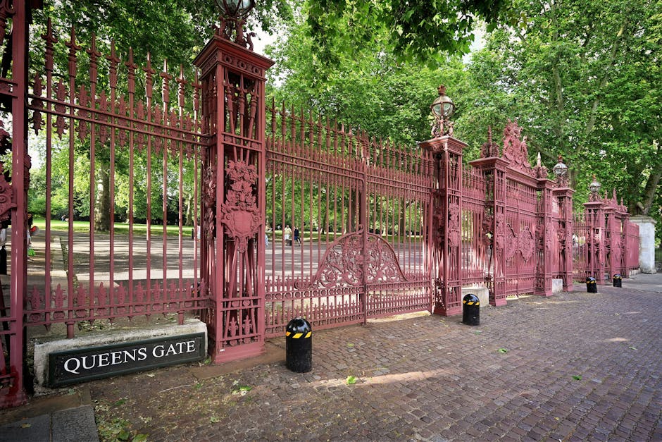 A large, ornate wrought iron gate painted in a weathered, mauve-red color, featuring intricate decorative elements and topped with multiple finials. The gate is supported by tall, square columns with 