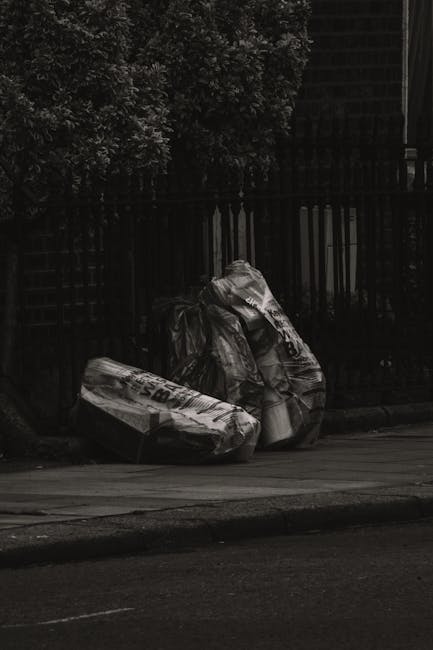 At the forefront of the image, there are multiple clear plastic bags filled with various types of discarded clothing, textiles, and miscellaneous waste, stacked on a paved sidewalk surface. The bags a